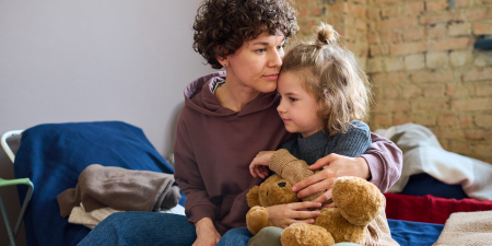 Young woman giving hug to her cute little son with brown soft teddybear while both sitting on sleeping place prepared for evacuees