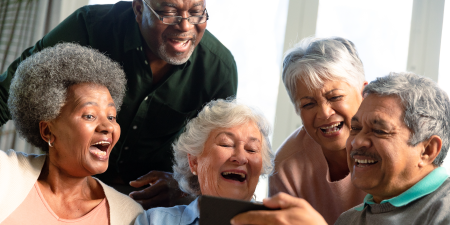 Five happy diverse senior friends sitting on sofa and looking at smartphone