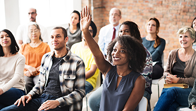 Image of a meeting with a woman in the front with her hand raised with a question.