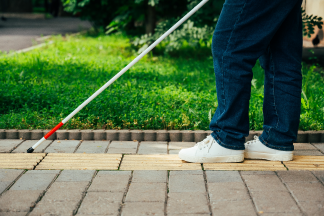 A blind woman walks outdoors using a cane along a tactile yellow tile.