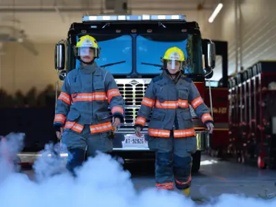 Two Clarington firefighters walking away from the fire truck in the background, white smoke rises from the ground around them.