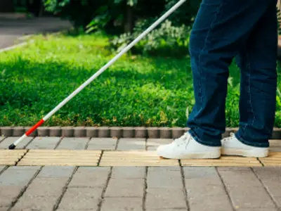 A blind woman walks outdoors using a cane along a tactile yellow tile.