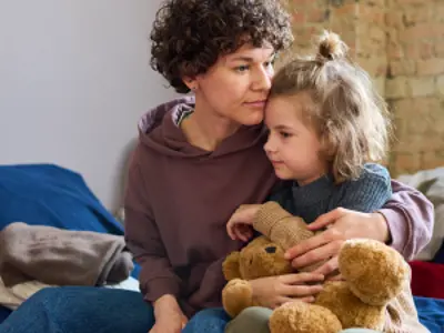 A parent holds a child at an emergency evacuation centre. They are sitting on cot and the child clutches a teddy bear.