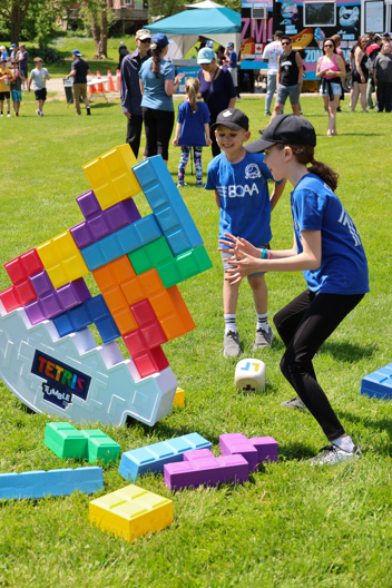 Children playing with a giant outdoor Tetris game, balancing blocks on a tipping base at the opening of Soper Creek Park Baseball Diamond.