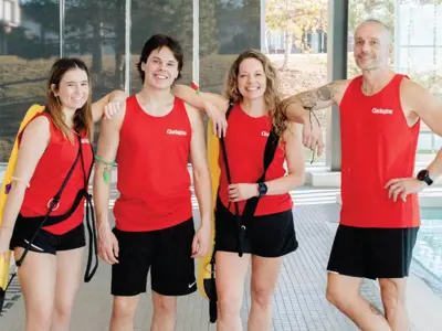 Four lifeguards in red tops and black shorts stand by an indoor pool, two holding yellow rescue tubes; large windows reveal trees outside.